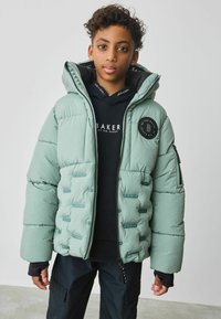 Padded jacket w kolorze miętowym z kapturem, z logo i kieszenią na zamek, noszony na czarnej bluzie z kapturem i ciemnych spodniach.