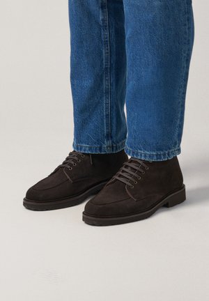 Bottines en daim marron avec un design à lacets, présentant un bout arrondi et une semelle en caoutchouc, associées à un jean en denim bleu.