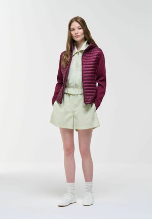 Young woman standing wearing light green zip-up jacket and shorts with a maroon quilted jacket, white socks, and white sneakers.