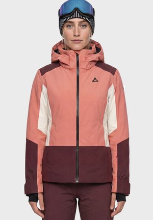 Frau trägt eine rosa, weiße und weinrote Skijacke mit Kapuze, eine schwarz-weiß gestreifte Mütze und blaue Skibrille auf der Stirn.
