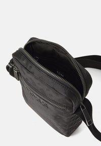 Sac bandoulière noir avec un extérieur texturé, une fermeture à double zip et un logo de marque. Bandoulière réglable et compartiment intérieur pour l'organisation.