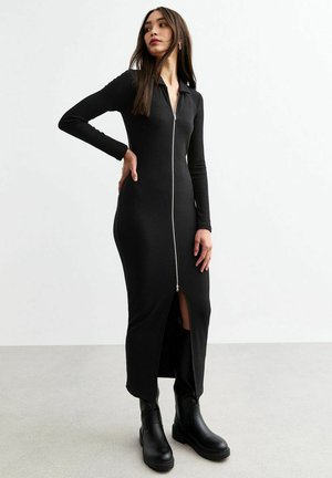 Robe noire à manches longues côtelées avec une fermeture éclair sur le devant, présentant une fente haute jusqu'à la cuisse et un ajustement près du corps. Portée avec des bottes noires épaisses.