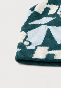 Pletená čiapka tmavozelenej farby s geometrickým vzorom v svetlomodrej a bielej. Textúrovaná látka s preloženým lemom na spodnej strane.