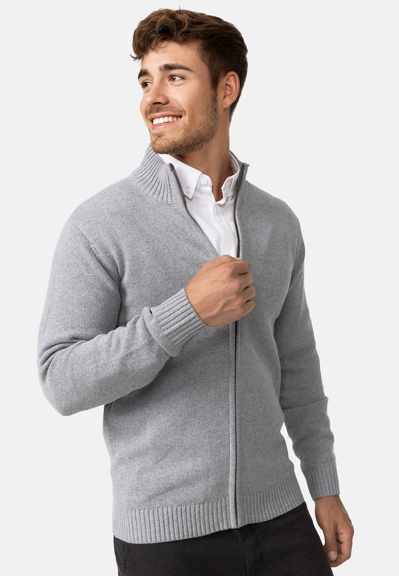 Sudadera gris con cremallera hecha de material de punto, con cuello y puños acanalados; presenta un corte recto y una textura suave. Combinada con una camisa blanca.