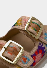 Sandales tissées avec une base beige naturelle, motifs brodés colorés en bleu, rose, jaune et vert, dotées de deux boucles en métal doré.