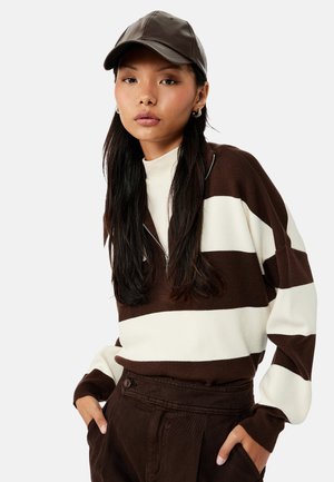 Vrouw met een bruin-wit gestreepte trui, een bruine broek en een donkerbruine leren baseballpet, poseert met handen in de zakken.