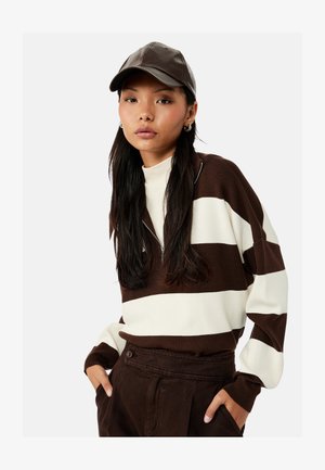 Frau trägt braun-weiß gestreiften Pullover, braune Hose und dunkelbraune Leder-Baseballkappe, posiert mit Händen in den Taschen.