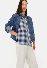 Denim jacket met knoopsluitingen, gecombineerd met een blauw-witte geruite shirt. Model draagt crèmekleurige broeken. Schone, minimalistische stijl.