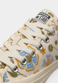 Baskets en toile Converse couleur blanc cassé avec broderie florale en bleu, vert, jaune et beige, dotées de lacets blancs et d'œillets métalliques.