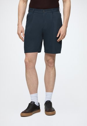 Uomo che indossa pantaloncini blu navy su misura, calzini bianchi e sneakers nere, in piedi davanti a uno sfondo semplice.