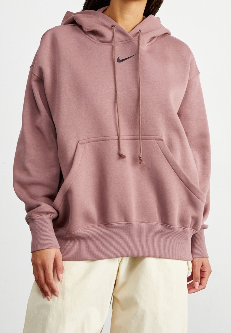 Hoodie oversize rosa con cordini, tasca anteriore, polsini e orlo a coste. Logo Nike nero centrato sul petto. Texture morbida.