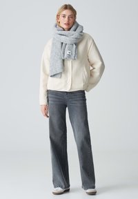 Beige bomberjack, grijs gebreid sjaaltje, donkergrijze flared jeans en lichtgrijze sneakers. Zachte texturen, ontspannen pasvorm en gelaagd ontwerp.