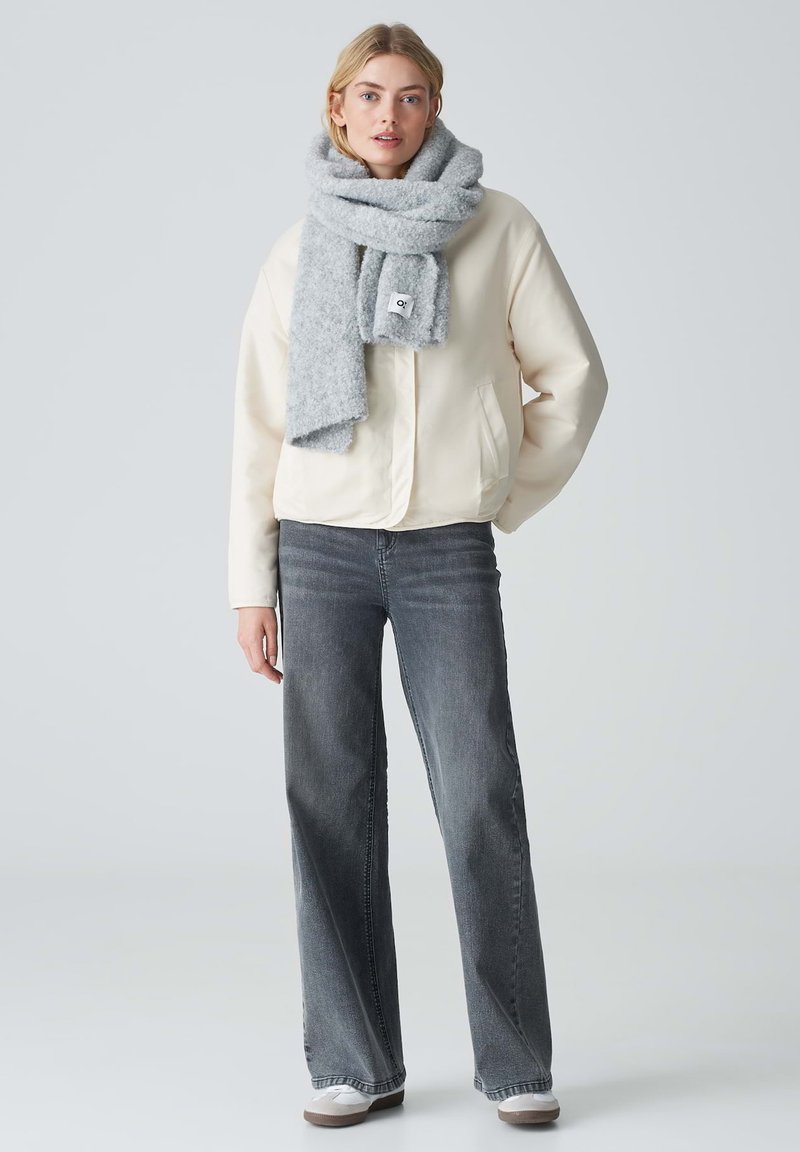 Beige bomberjack, grijs gebreid sjaaltje, donkergrijze flared jeans en lichtgrijze sneakers. Zachte texturen, ontspannen pasvorm en gelaagd ontwerp.