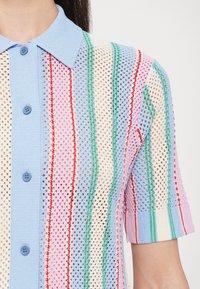 Gros plan d'un pull à manches courtes avec col bleu et rayures verticales multicolores en rose, vert, rouge et crème sur un motif en maille.