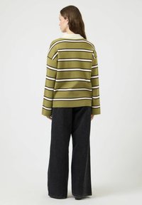 Kvinne med langt brunt hår iført en olivengrønn stripete genser og svarte vide bukser, stående med ryggen mot mot en ensfarget bakgrunn.