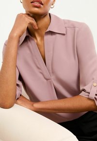 Blusa di un delicato colore malva con scollatura a V, maniche corte arrotolate e dettagli con bottoni dorati. Tessuto liscio con un taglio aderente.