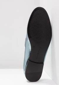 Chaussure bleu métallique à finition lisse, semelle extérieure plate noire avec un motif texturé, étiquette visible sur la semelle indiquant la taille 37.