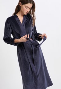 Femme portant une robe en velours bleu foncé, nouant la ceinture à la taille avec les deux mains, regardant vers le bas, sur un fond blanc uni.