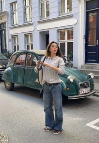 Žena v sivom svetri a džínsoch držiaca slnečné okuliare stojí vedľa retro zeleného auta na mestskej ulici s budovami s modrými dverami.