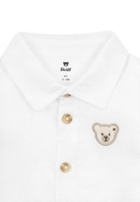 Camisa polo blanca con tejido texturizado, tres botones dorados y una cara de oso bordada en el lado izquierdo, etiqueta de la marca en el interior del cuello.