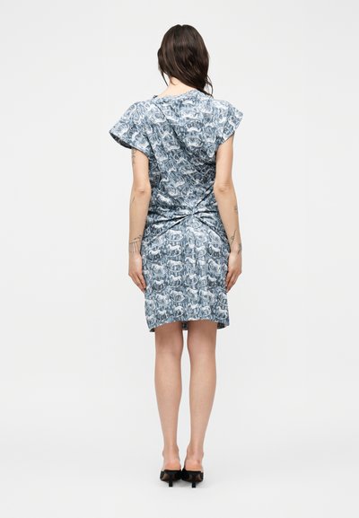 Γυναίκα με σκούρα μαλλιά που φοράει μπλε φόρεμα με μοτίβα, κοντομάνικο και μήκους μέχρι το γόνατο, και μαύρα ψηλοτάκουνα, να στέκεται με την πλάτη προς το μέρος μας σε απλό φόντο.