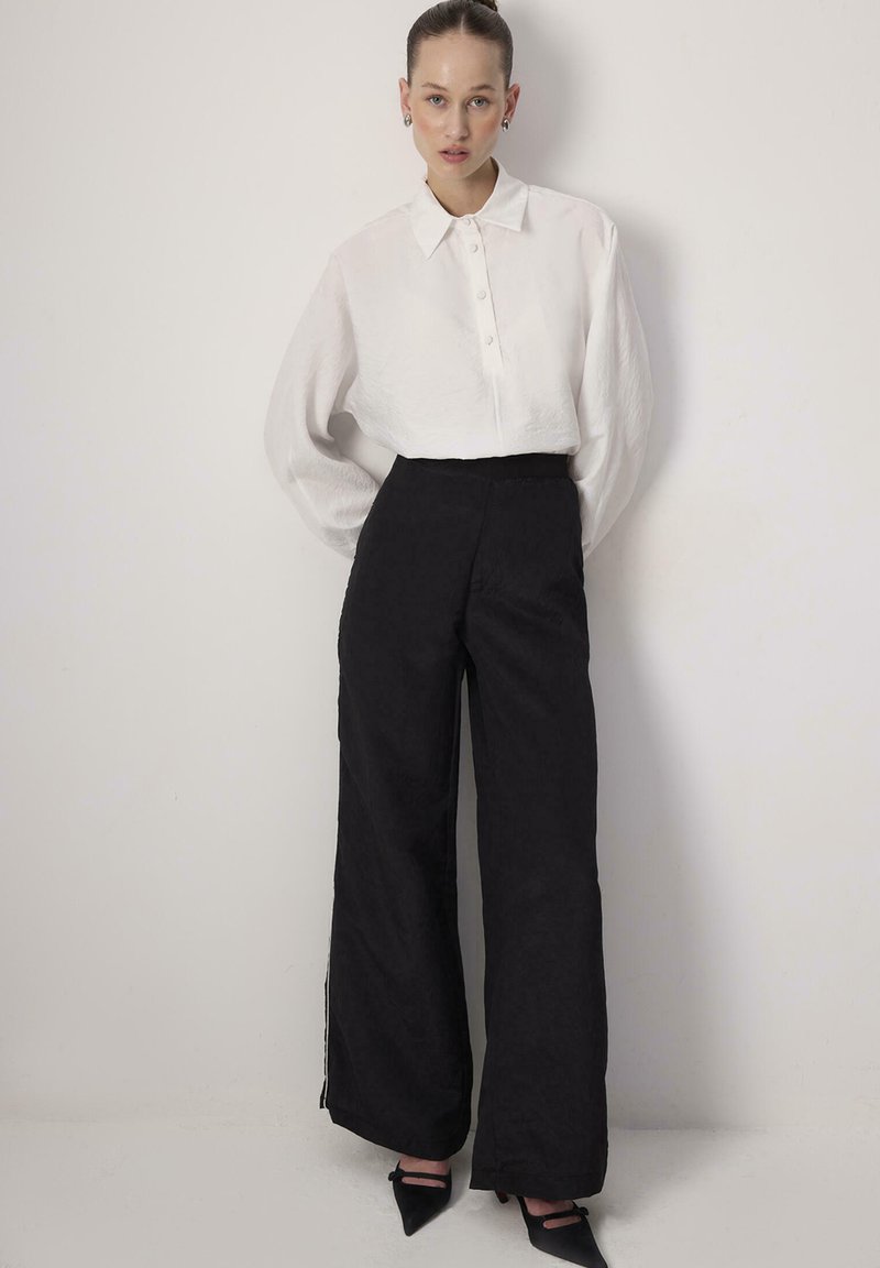 Γυναίκα που στέκεται μπροστά σε λευκό τοίχο, φοράει λευκό πουκάμισο με κουμπιά και μακρύ μανίκι, μαύρο παντελόνι με φαρδιά μπατζάκια και ψηλοτάκουνα μαύρα παπούτσια.