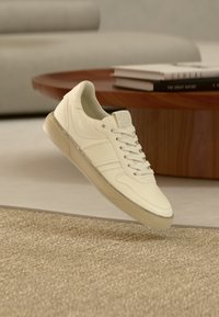 Λευκό δερμάτινο αθλητικό παπούτσι με κορδόνια, υφασμάτινες λεπτομέρειες και διαφανές καουτσούκ σόλα, παρουσιασμένο πάνω σε υφαντή επιφάνεια.