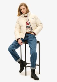 Creme pufferjakke med trykknapper, båret over en stribet sweater. Blå denimjeans med opsmøg i buksebenene og sorte snørebåndsstøvler. Sidder på en skammel.