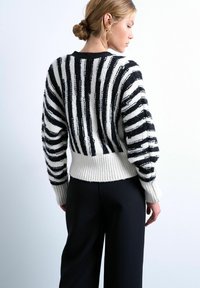 Femme aux cheveux blonds en chignon, portant un pull en tricot rayé noir et blanc et un pantalon noir, vue de profil sur un fond uni.