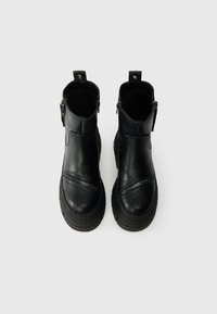 Par de botas de tornozelo em couro preto com fechos laterais e solas de borracha grossa, vistas de cima sobre um fundo branco.