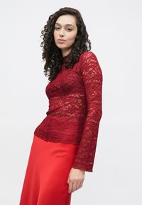 Rdeča čipkasta majica z dolgimi rokavi, okrašena z cvetličnim vzorcem in valovitim robom. V kombinaciji s trdo rdečo krilo.