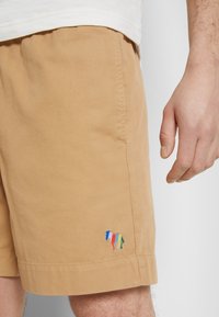 Pantalones cortos color beige, confeccionados en una mezcla de algodón, con un colorido diseño bordado de un caballo en la pierna izquierda. Textura suave, corte recto y dobladillo enrollado.