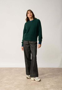 Zielona dziana bluza z zamkiem i niebieskimi dzianinowymi mankietami, w zestawieniu z ciemnoniebieskimi spodniami typu wide-leg i jasnymi sneakersami. Gładkie tło.