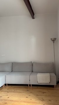 Γκρίζος γωνιακός καναπές με τρία μαξιλάρια και διπλωμένη μπεζ ριχτάρι πάνω σε ξύλινο πάτωμα, δίπλα σε έναν ψηλό ασημένιο επιδαπέδιο φωτιστικό.