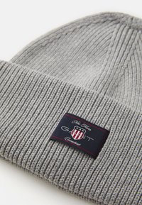 Beanie en tricot gris à côtes avec un revers et un patch logo rectangulaire portant le texte "GANT", un écusson et des rayures rouges et bleues.