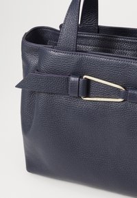 Navy kožená kabelka s texturovaným povrchem. Má ozdobný přezkový prvek a dvojité držadla, které zdůrazňují strukturovaný design s elegantním tvarem.