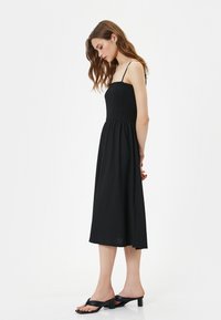 Schwarzes Sommerkleid mit gerafftem Oberteil und dünnen Trägern, das bis zur Wadenmitte reicht. Kombiniert mit schwarzen High Heels Sandalen. Sanfter, leichter Stoff.