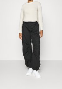 Schwarze Cargo-Hosen mit lockerer Passform und elastischen Bündchen an den Knöcheln, kombiniert mit einem cremefarbenen gerippten Pullover. Weiße Sneaker runden das Outfit ab.