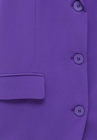 Veste violette en tissu lisse, avec un revers cranté et quatre boutons. Les boutons sont placés à l'avant, ronds et ont une finition mate.