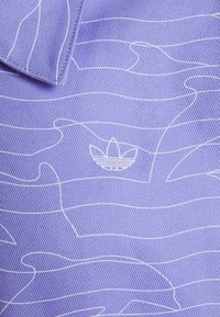 Lila texturerad tyg med vitt vågmönster, med en liten, broderad Adidas-logotyp i det nedre vänstra hörnet. Standard polo kragsdesign.