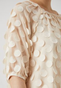 Sheer, beige blouse featuring circular fabric appliqués and elasticized puff sleeves. The texture is light and slightly layered with a soft finish.