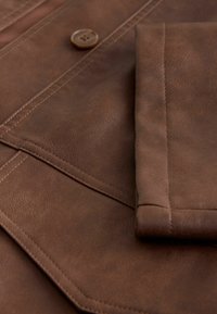Gros plan sur la manche d'une veste en cuir marron, rabat de poche, couture visible et un unique bouton marron sur un tissu en cuir texturé.