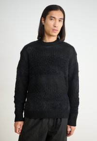 Black knitted sweater featuring textured patterns, varying weaves, and a ribbed neckline. Long sleeves with a relaxed fit.