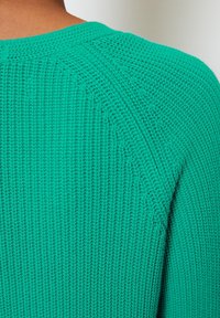 Pull en tricot vert avec un motif texturé, présentant un col rond et des manches raglan. Le tissu semble léger et respirant.