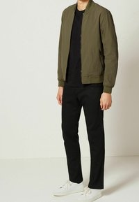 Olivenfarbene Bomberjacke mit Reißverschluss, gerippten Bündchen und zwei seitlichen Taschen, getragen über einem schwarzen Hemd und schwarzen Hosen, kombiniert mit weißen Sneakers.