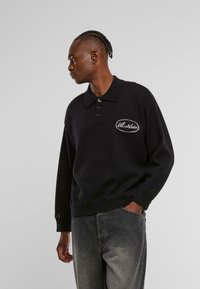 Schwarzer Strick-Polo-Pullover mit langen Ärmeln, dreiknöpfiger Knopfleiste und gesticktem ovalen Logo auf der linken Brust, kombiniert mit dunkelgrauen Jeans.