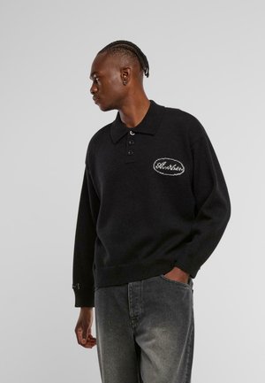 Maglione polo a maniche lunghe nero in maglia con chiusura a tre bottoni e logo ovale ricamato sul lato sinistro del petto, abbinato a jeans grigio scuro.