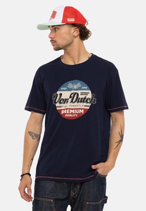 Homme portant un t-shirt bleu marine Von Dutch et une casquette Von Dutch rouge et blanche, regardant vers la gauche, avec des tatouages de papillon et d'araignée sur les bras.