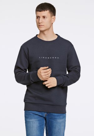 Sweatshirt blu navy realizzato in cotone misto, con colletto a girocollo e polsini a costine. Testo del logo "LINDGERGH" centrato sulla parte anteriore. Indossato con jeans blu.