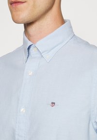 Homme portant une chemise bleu clair à boutons avec col boutonné et un petit logo brodé sur la poitrine.
